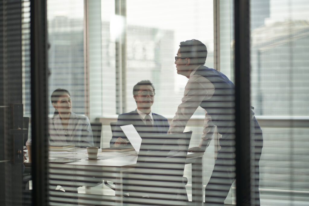 through-the-glass shot of an asian manager standing at the top of meeting table speaking team members