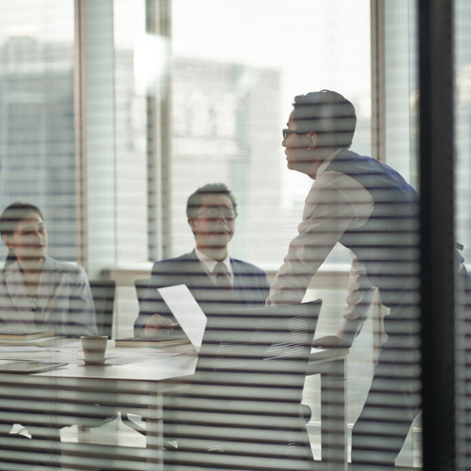 through-the-glass shot of an asian manager standing at the top of meeting table speaking team members